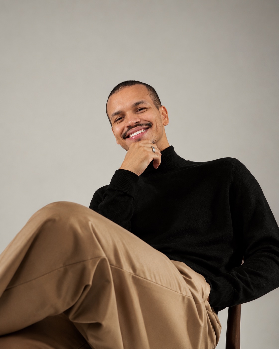 Ryan Alves sorrindo, de blusa preta de gola alta, sentado, com a mão no queixo